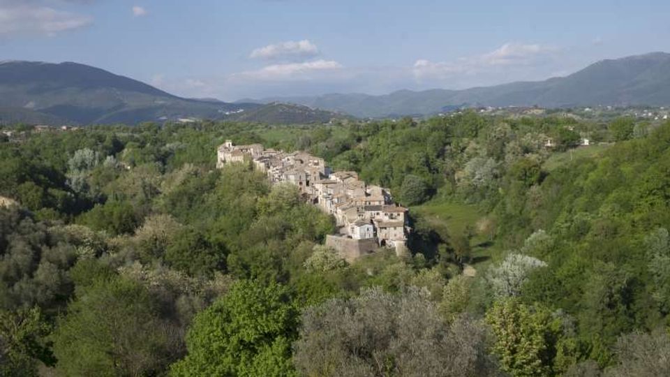Veduta panoramica di un piccolo borgo con case in pietra addossate su un crinale, circondato da una fitta vegetazione. Sullo sfondo si vedono colline e montagne sotto un cielo parzialmente nuvoloso.