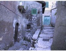 Vicolo stretto in pietra con gradini che salgono tra case antiche intonacate e muri invecchiati. Si vedono una porta ad arco, piccole finestre con persiane verdi e una grande porta verde in fondo al passaggio.