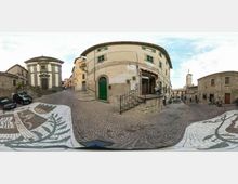 Piazza lastricata in pietra con un grande mosaico decorativo sul pavimento, circondata da edifici storici in muratura e una piccola chiesa con scalinata. Al centro si vede un edificio curvo con porte verdi e una scala con ringhiera, mentre la piazza si apre verso vie strette del borgo.
