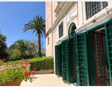 Facciata di un edificio chiaro con ingresso ad arco e grandi persiane verdi aperte, affacciato su un cortile pavimentato. Accanto all’edificio si vede un giardino con siepi, fiori e alte palme sotto un cielo sereno.