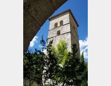 Torre campanaria in pietra con tetto a falde e aperture ad arco, vista dal basso tra alberi e vegetazione. Sullo sfondo si vede il cielo azzurro con alcune nuvole e una collina boscosa.