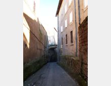 Vicolo stretto in pendenza tra alti edifici in muratura intonacata, con finestre sbarrate e tubazioni esterne lungo le pareti. In fondo si vede un piccolo ponte ad arco in pietra sopra la strada, con muschio e segni di umidità sulle basi dei muri.