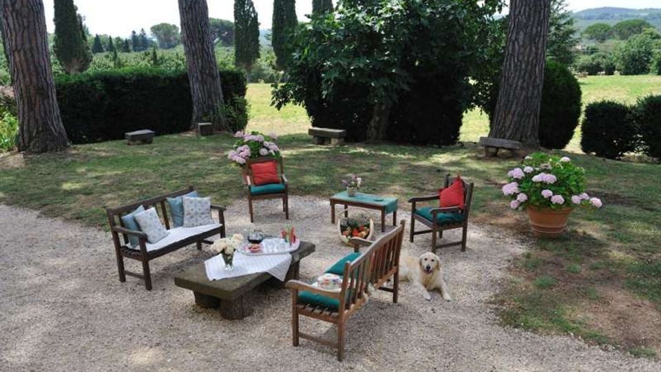 Area salotto all’aperto su ghiaia in un giardino, con sedute in legno e tavolini attorno a un tavolo con tovaglia. Intorno ci sono grandi pini, siepi e vasi di ortensie, con vista su un prato e colline sullo sfondo.