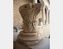 Capitello in pietra scolpita con motivi vegetali e figure stilizzate, appoggiato su una base circolare. Sullo sfondo si vede un porticato in muratura con colonne e altri capitelli esposti lungo il corridoio.
