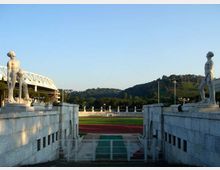 Ingresso monumentale di uno stadio con due statue in pietra ai lati e gradinate in marmo che conducono a una pista di atletica rossa e al campo centrale. Sullo sfondo si vedono colline verdi e una tribuna con struttura metallica sotto un cielo sereno.
