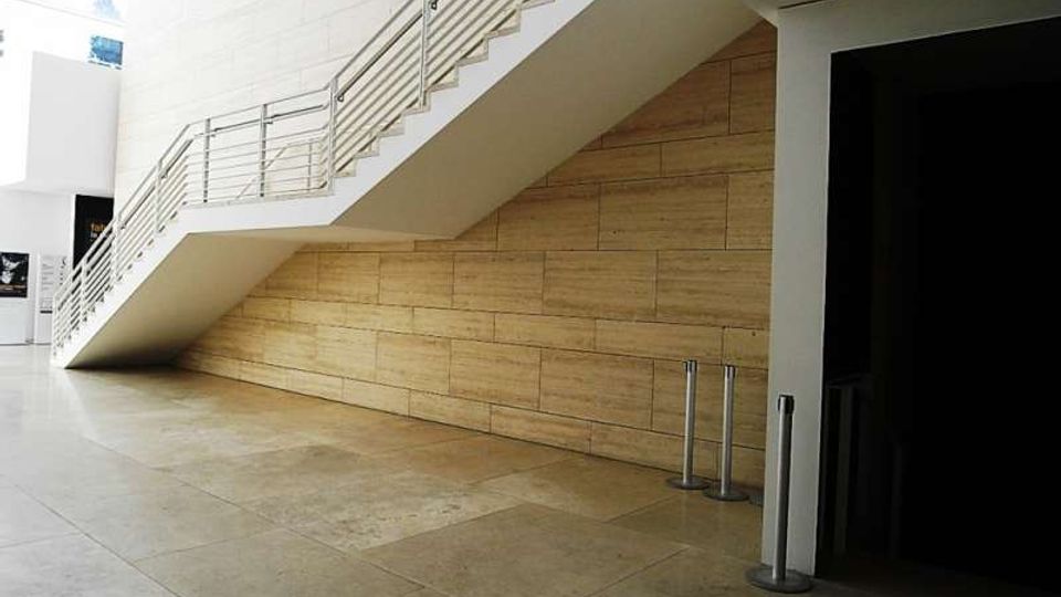 Atrio interno moderno con ampia scalinata bianca e corrimano in metallo che sale lungo una parete rivestita in pannelli di legno. Pavimento in grandi lastre di pietra chiara e ingresso scuro laterale con colonnine metalliche.