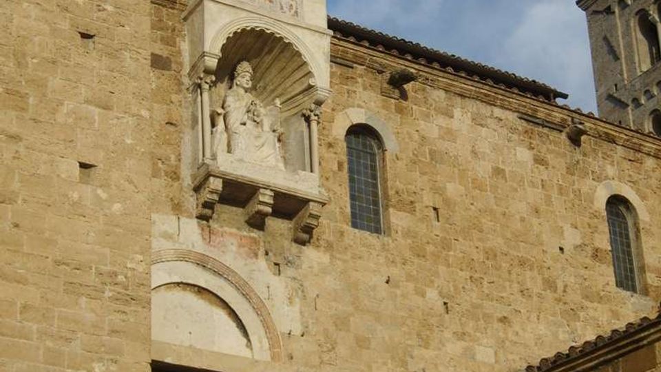 Facciata esterna di un edificio storico in pietra, con finestre ad arco e un portale sormontato da una nicchia sporgente con scultura e baldacchino decorato. In primo piano si vede una terrazza con ringhiera in ferro e, sullo sfondo, un tratto di torre e cielo.
