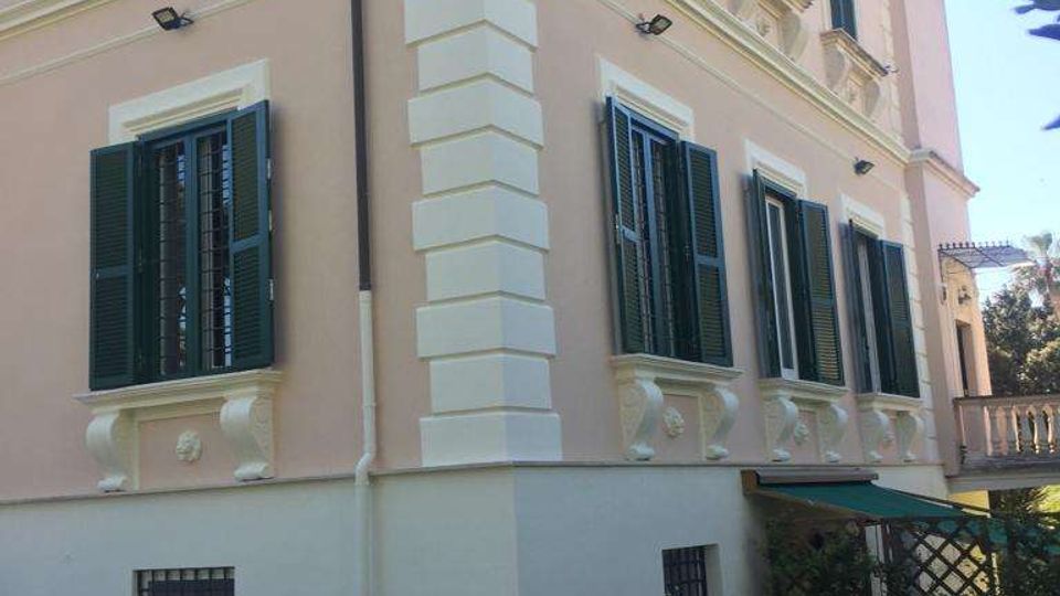 Facciata angolare di una villa a più piani intonacata in rosa con cornici bianche, persiane verdi e finestre alte con dettagli decorativi. In primo piano si vedono un vialetto in pietra con motivo circolare, un prato e un vaso su piedistallo vicino a un’area con pergolato.