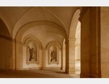 Interno di un porticato in pietra con volte a botte e archi, sostenuto da pilastri massicci. Sulla parete di fondo si vedono due nicchie ad arco con statue e pavimento in pietra.