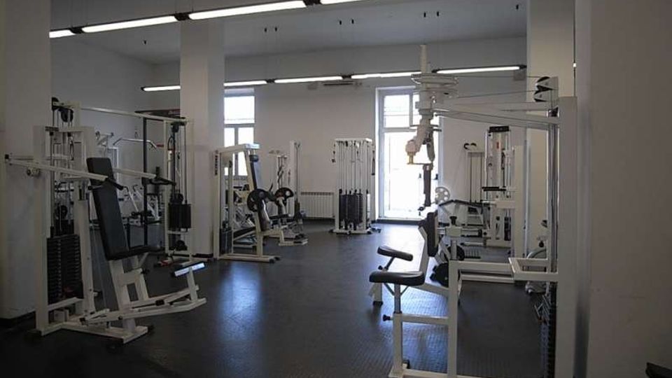 Ampia sala palestra interna con pavimento scuro in gomma, pareti bianche e file di macchine per pesi e cavi disposte lungo i lati. Il locale è illuminato da lunghe lampade fluorescenti a soffitto e da grandi finestre sul fondo.