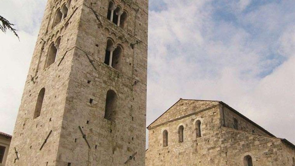 Torre campanaria medievale in pietra con alte bifore sovrapposte e grandi arcate alla base, accanto a una chiesa romanica in pietra con facciata semplice e portale ad arco. Inquadratura dal basso con cielo parzialmente nuvoloso, rami di albero e un lampione in alto a destra.