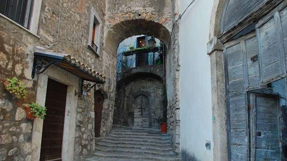 Vicolo in pietra in un borgo storico con una scalinata centrale che sale sotto archi in muratura. Ai lati si vedono facciate in pietra e intonaco, porte in legno e un piccolo balcone con ringhiera e piante.