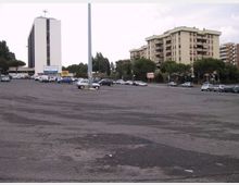 Ampio parcheggio asfaltato quasi vuoto, con diverse auto parcheggiate e un alto palo della luce al centro. Sullo sfondo si vedono un edificio moderno a torre bianco e vari palazzi residenziali con balconi, circondati da alberi lungo la strada.