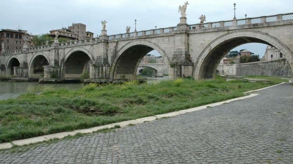 Ponte storico in pietra con più arcate che attraversa un fiume, con statue lungo la balaustra e lampioni. In primo piano si vede una strada acciottolata con una fascia erbosa lungo la riva, e sullo sfondo edifici urbani.