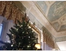 Interno di una sala con soffitto alto decorato a riquadri con stucchi e motivi classici, cornici ornate e tende pesanti a fantasia. In primo piano si vede un albero di Natale illuminato con lucine, vicino a una finestra e a uno specchio con cornice dorata.