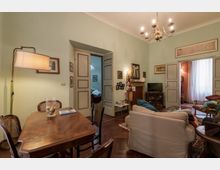 Interno di un soggiorno-sala da pranzo con pareti verde chiaro, pavimento in parquet e lampadario a bracci sul soffitto. Al centro ci sono un tavolo in legno con sedie, un divano chiaro e un’area TV vicino a un camino, con porte a pannelli grigi che conducono ad altre stanze illuminate.