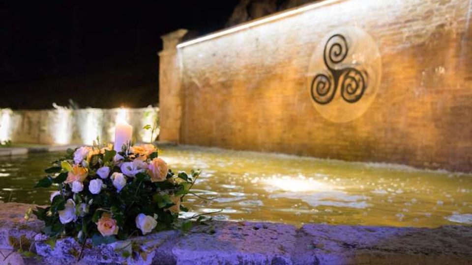 Fontana notturna con vasca d’acqua illuminata e parete in pietra sullo sfondo, decorata da un grande simbolo a spirale. In primo piano sul bordo in pietra c’è un mazzo di fiori con una candela accesa.