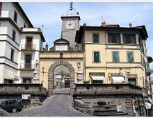 Piazza cittadina con rampa in pavé che conduce a un grande arco in pietra inserito in un edificio storico, con torre dell’orologio sullo sfondo. Intorno si vedono palazzi a più piani con persiane verdi e insegne di negozi, oltre a muretti in pietra e una piccola fontana in primo piano.