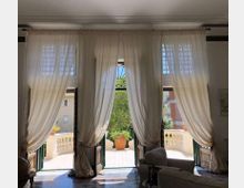 Interno di una stanza con tre alte porte-finestre affiancate, coperte da tende bianche leggere, che si aprono su una terrazza con balaustra in pietra. All’esterno si vede un grande vaso al centro e la vegetazione, con edifici sullo sfondo.