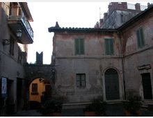 Cortile interno in un centro storico, circondato da edifici in intonaco consumato con persiane verdi e tetti in coppi. A sinistra si vede un arco in pietra che conduce a un passaggio illuminato, con porte e finestre ad arco e vasi di piante lungo il pavimento in pietra.