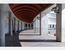 Portico coperto lungo e curvilineo con arcate e soffitto in legno, sostenuto da colonne e pilastri rivestiti in marmo con lampade a parete. Pavimentazione in pietra con motivi circolari e vista laterale su una strada con auto parcheggiate e un edificio moderno sullo sfondo.