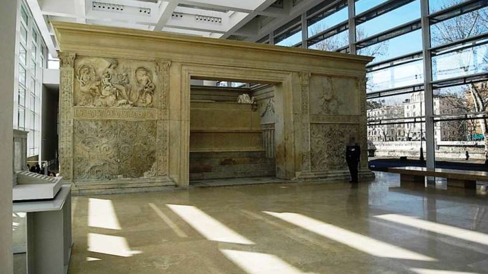 Interno di un museo o atrio moderno con ampie vetrate a tutta altezza e soffitto a griglia, che ospita una grande struttura in pietra scolpita con bassorilievi e un’apertura centrale. Pavimento lucido in pietra con fasci di luce e ombre proiettate dalle finestre.