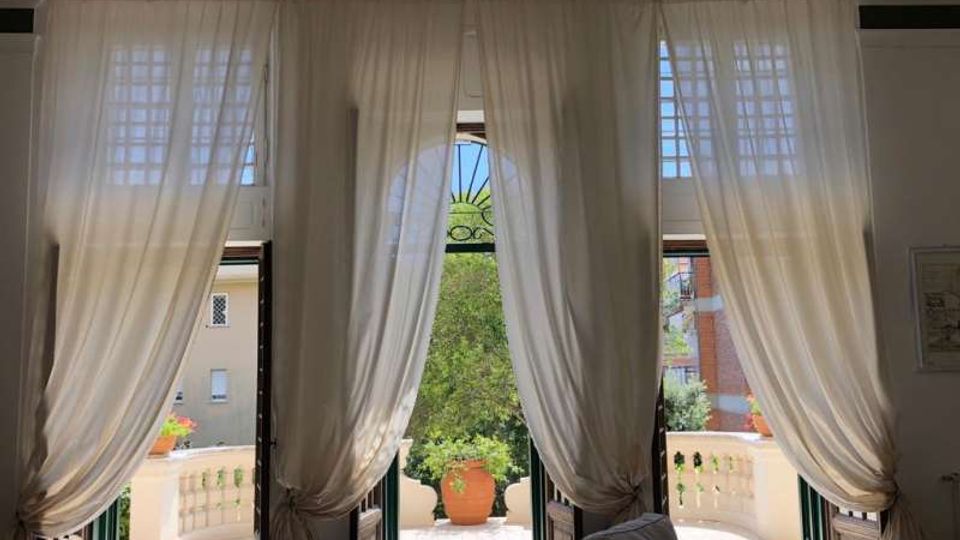 Interno di una stanza con tre alte porte-finestre affiancate, coperte da tende bianche leggere, che si aprono su una terrazza con balaustra in pietra. All’esterno si vede un grande vaso al centro e la vegetazione, con edifici sullo sfondo.