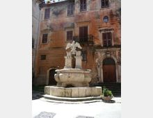 Fontana in pietra al centro di una piccola piazza, con vasca circolare su gradoni e scultura di un’aquila su piedistalli. Sullo sfondo si vede la facciata di un edificio storico intonacato, con persiane scure, balconcino in ferro e portale ad arco decorato.