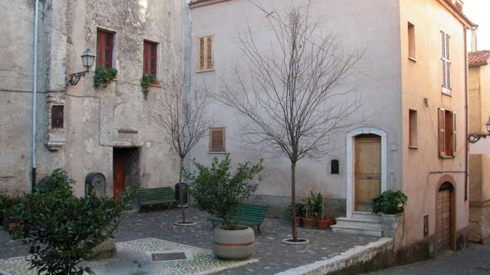 Piccola piazzetta lastricata in un borgo, con panchine verdi, vasi con arbusti e due alberi spogli. Intorno si affacciano case in muratura con intonaco chiaro, finestre con persiane e una porta ad arco sopra alcuni gradini.