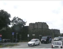 Strada urbana con traffico veicolare in primo piano, affiancata da una grande fortificazione in pietra con mura spesse e torre circolare. Sullo sfondo si vedono alberi e segnaletica stradale sotto un cielo nuvoloso.