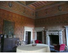 Interno di una sala storica con pareti affrescate e soffitto a cassettoni decorato, dotata di grande camino in pietra con cornice scolpita e porte con stipiti bianchi. Al centro c’è un tavolo rotondo coperto da tovaglia, con sedie e poltrone rosse, e un grande dipinto appeso sopra una boiserie.