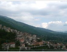 Veduta panoramica di un borgo collinare con case in pietra e tetti in tegole addossate al pendio, ai piedi di una catena montuosa. Sullo sfondo si estende una valle verde con campi e boschi sotto un cielo nuvoloso.
