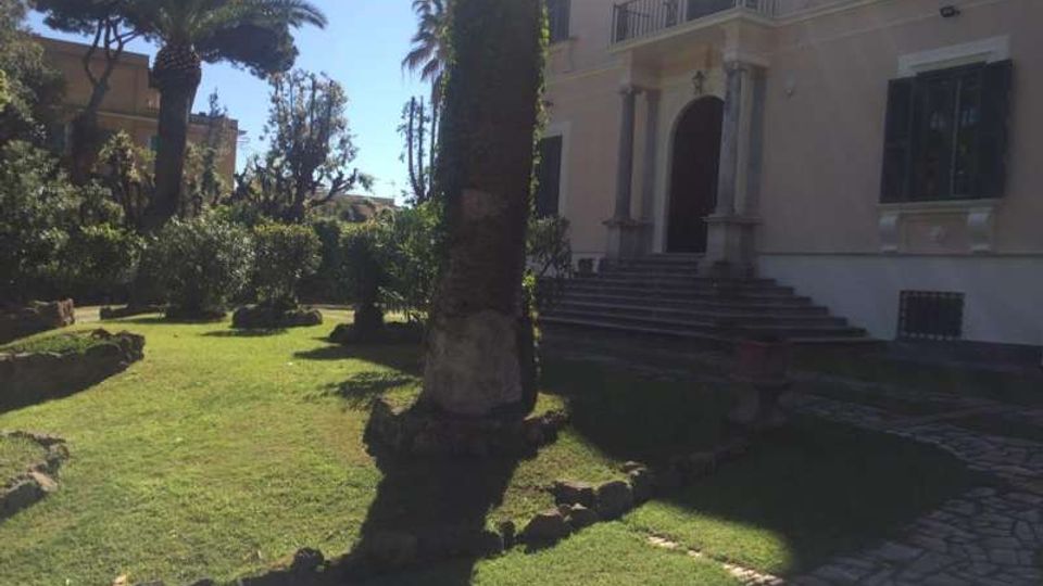 Giardino con prato curato, palme e arbusti, attraversato da vialetti in pietra e bordi in mattoni. Sullo sfondo si vede una villa chiara con ingresso ad arco sorretto da colonne e una scalinata che porta al portone.