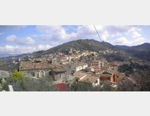 Veduta panoramica di un borgo collinare con case in pietra e tetti in coppi, disposto su pendii terrazzati. Sullo sfondo si vedono colline boscose e un cielo parzialmente nuvoloso.