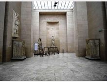 Interno di una grande sala museale in pietra chiara, con pavimento in lastre e alto lucernario a soffitto. Al centro una statua su piedistallo e attrezzature sceniche in legno, affiancate da due grandi basamenti scolpiti e una nicchia sul fondo.