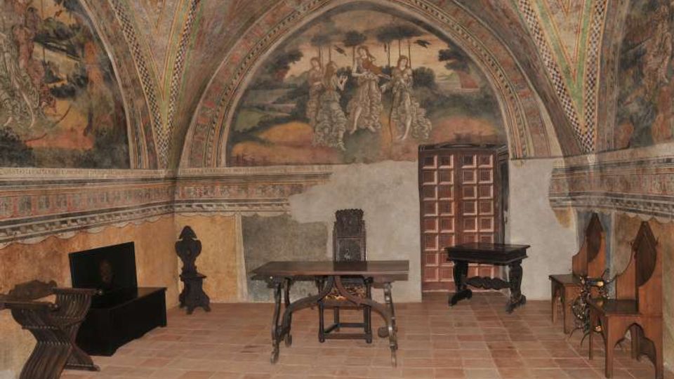 Interno di una sala storica con soffitto a volta affrescato e lunette dipinte, pareti decorate e pavimento in cotto. Al centro ci sono tavoli e sedie in legno, con una porta a pannelli sul fondo e arredi scuri lungo le pareti.