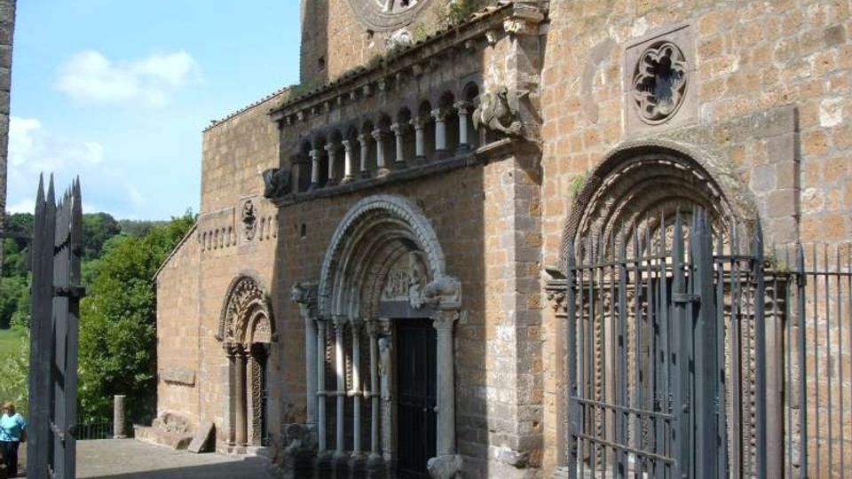 Facciata in pietra di una chiesa medievale con portali ad arco decorati, loggetta con colonnine e grande rosone sopra l’ingresso principale. Davanti si vede un sagrato in pietra con cancellate metalliche e, sullo sfondo, alberi e cielo.