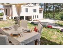Tavolo da pranzo apparecchiato sotto un gazebo con tende bianche, in un giardino davanti a una villa chiara con persiane grigie e ampie finestre. Sullo sfondo si vedono un vialetto in ghiaia, aiuole fiorite e alberi che circondano la proprietà.