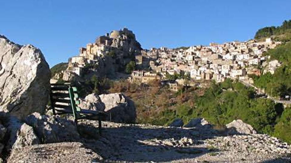 Veduta panoramica di un borgo arroccato su una collina rocciosa, con case in pietra addossate l’una all’altra e un edificio fortificato in cima. In primo piano si vedono un terreno ghiaioso con grandi massi e una panchina, circondati da vegetazione e cielo sereno.