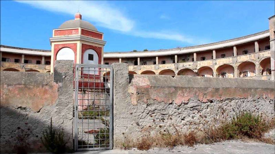 Cortile interno di una struttura fortificata con porticati ad archi su due livelli e pareti intonacate. In primo piano un basso muro in pietra e un cancello metallico, sullo sfondo una piccola torretta ottagonale rossa e bianca con cupola sotto un cielo azzurro.