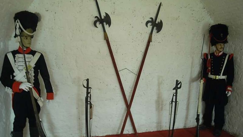Interno di una nicchia ad arco con pareti bianche e pavimento rosso, allestita come esposizione storica con armi in asta incrociate al centro. Ai lati sono presenti due manichini in uniforme d’epoca e vari fucili e sciabole esposti su supporti.