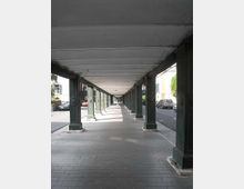 Portico urbano lungo e rettilineo con pavimentazione in pietra e soffitto a travi, sostenuto da una fila di colonne in metallo scuro. Ai lati si intravedono la strada con auto parcheggiate e alcuni edifici.