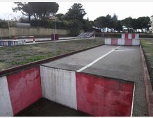 Pista asfaltata all’aperto con cordoli e barriere basse dipinte di rosso e bianco, disposta in corsie e curve come un circuito di guida. Sullo sfondo si vedono un muro di recinzione, lampioni e alberi alti lungo il perimetro.