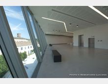 Ampio interno di edificio moderno con pavimento chiaro, soffitto con illuminazione lineare e grandi vetrate inclinate a tutta altezza sul lato sinistro. Dalle finestre si vede un complesso storico con cortile e campanile; lungo la sala sono presenti una panca scura e pareti bianche con porte.