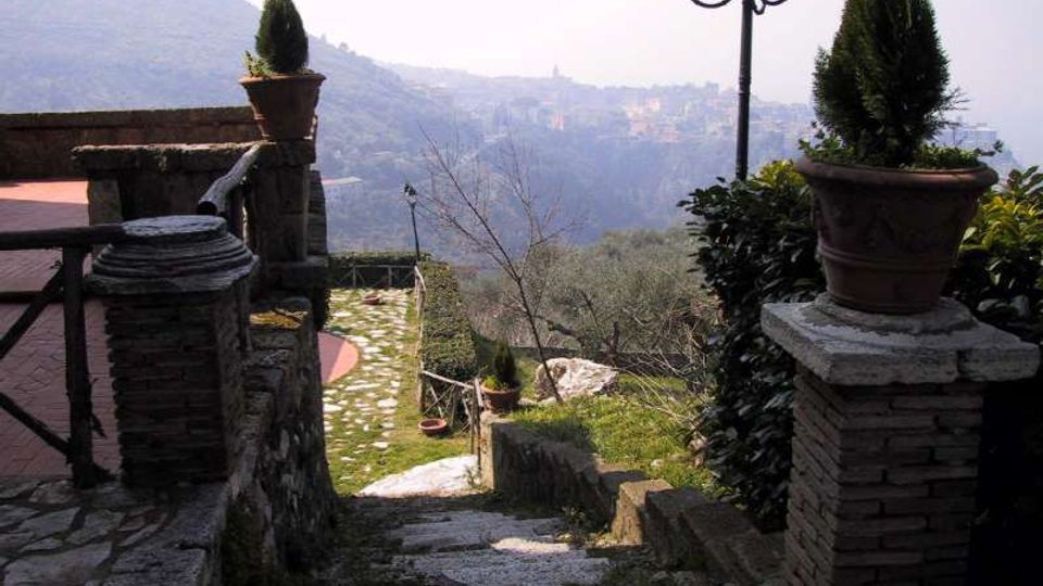 Scalinata in pietra che scende da una terrazza con muretti in muratura e vasi con arbusti, affiancata da un lampione a più luci. Sullo sfondo si vede una valle con uliveti e un borgo collinare in lontananza.