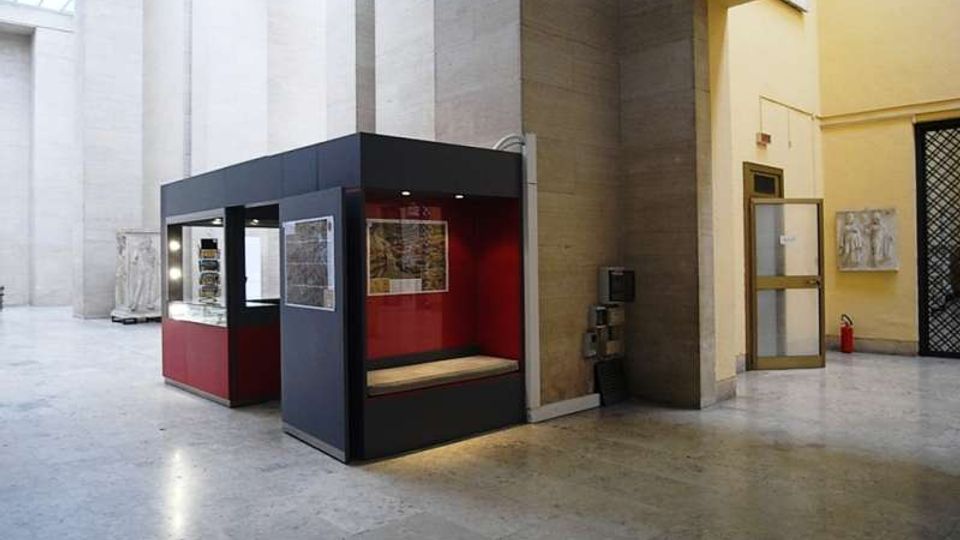 Ampio atrio interno con pavimento in pietra e alte pareti in cemento chiaro illuminate da lucernari. Al centro è presente un chiosco espositivo moderno nero e rosso con vetrine e pannelli, accanto a un passaggio verso un corridoio giallo con porte e grate.