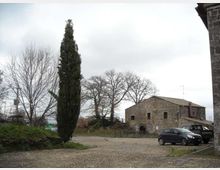 Piazzale lastricato in pietra con un alto cipresso e alberi spogli, affacciato su un edificio rurale in pietra con tetto a falde. Sullo sfondo cielo nuvoloso e alcune auto parcheggiate lungo il bordo della piazza.