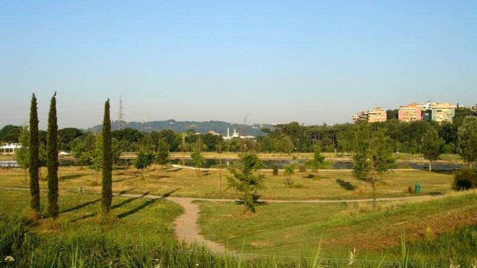 Ampio parco urbano con prati ondulati e sentieri sterrati, alberi sparsi e alcuni cipressi slanciati in primo piano. Sullo sfondo si vedono un corso d’acqua o canale, colline e palazzi residenziali sotto un cielo sereno.