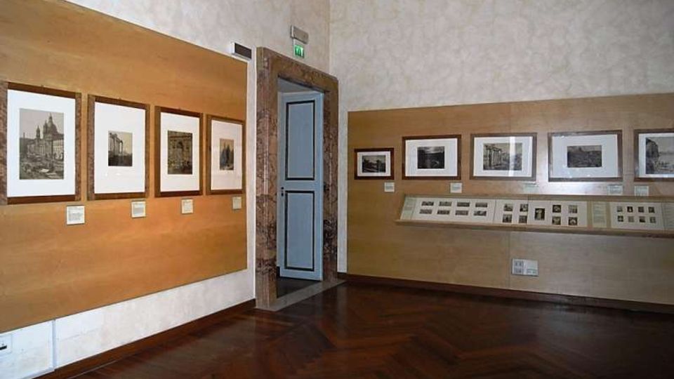 Interno di una sala espositiva con pareti chiare e pannelli in legno che ospitano fotografie incorniciate e didascalie. Pavimento in parquet scuro e porta centrale con cornice in pietra, con una lunga vetrina orizzontale a parete che espone materiali e immagini.