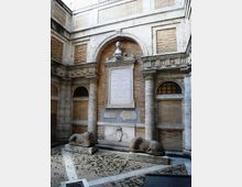Cortile interno in stile classico con pareti in mattoni e cornici in pietra, archi e colonne laterali. Al centro c’è una lapide commemorativa incassata sotto un arco con busto scolpito, affiancata da due leoni in pietra sdraiati su basamenti su pavimentazione a mosaico.